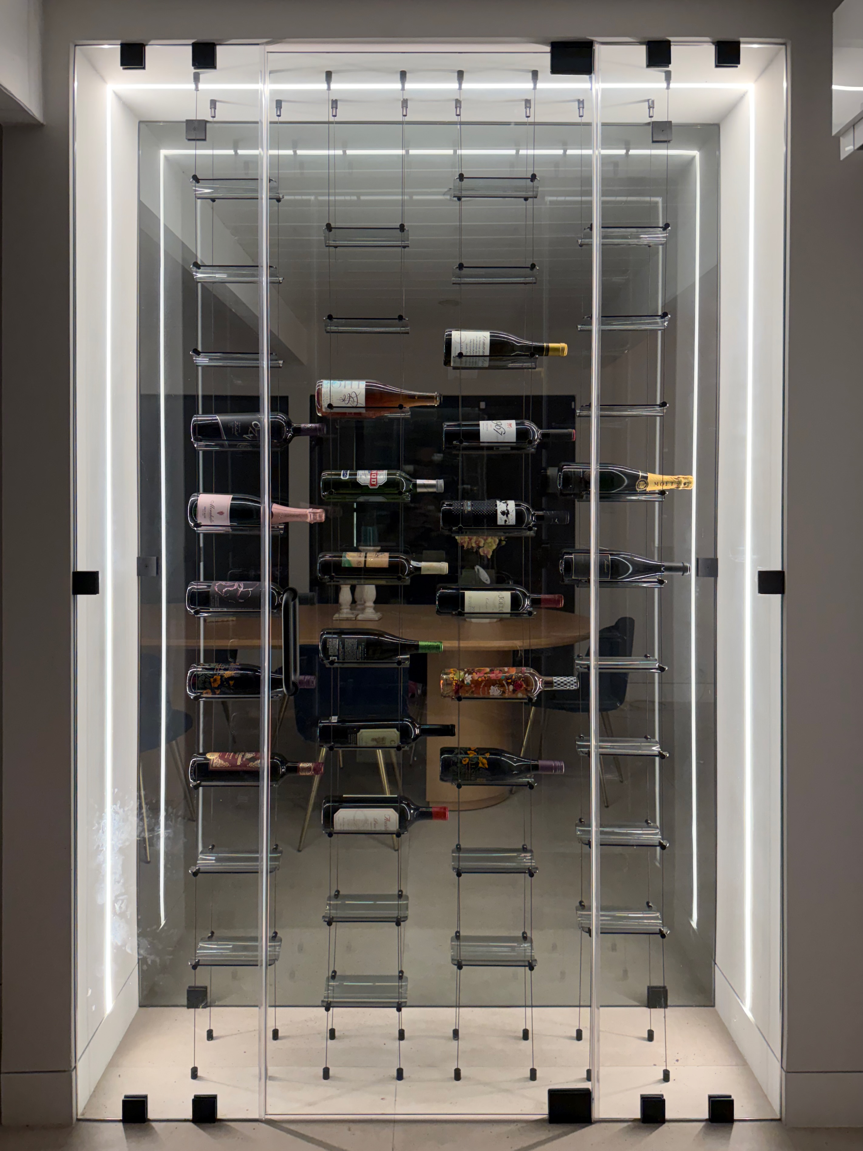 Symmetrical full-frontal view of a modern glass wine gallery wall with floating wine bottles suspended on stainless steel cable racks inside a frameless glass enclosure with integrated LED lighting