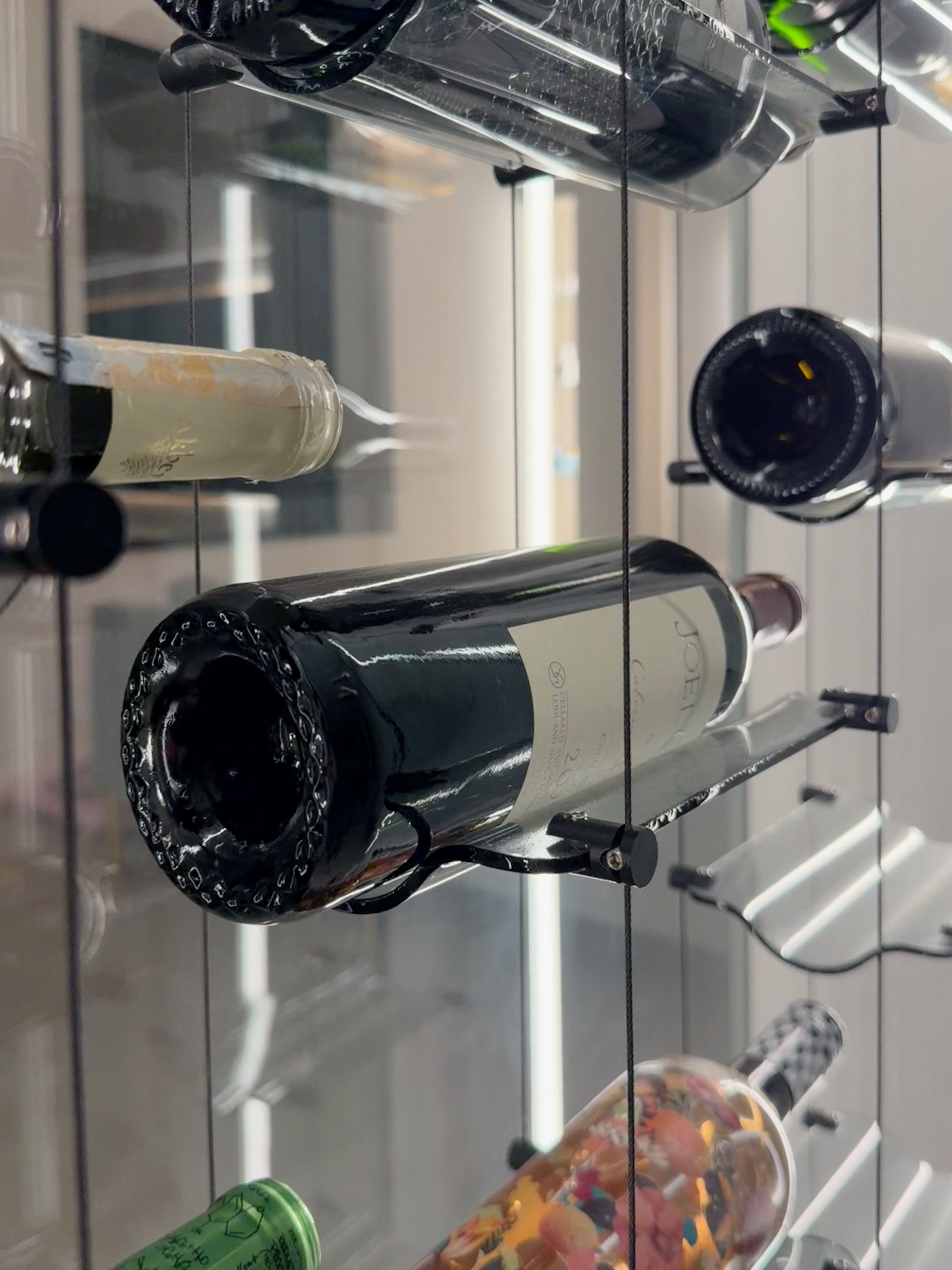 Close-up photograph of a wine bottle resting on a transparent glass rack mounted to a stainless steel cable support system with minimal hardware